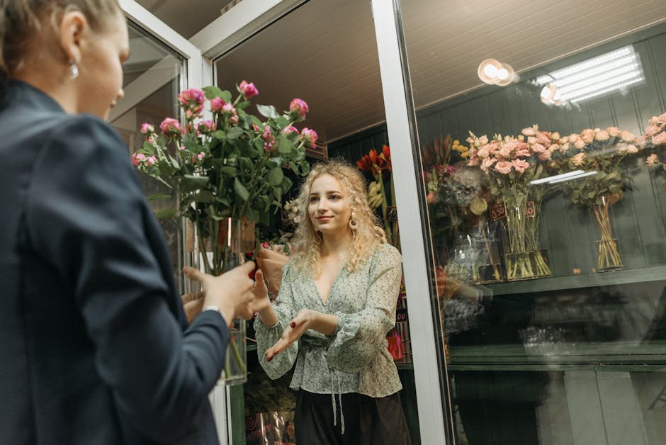 A young woman with curly blonde hair and wearing a light-colored, patterned blouse accepts a bouquet of freshly cut pink roses wrapped in brown paper from a florist inside a flower shop. The florist,