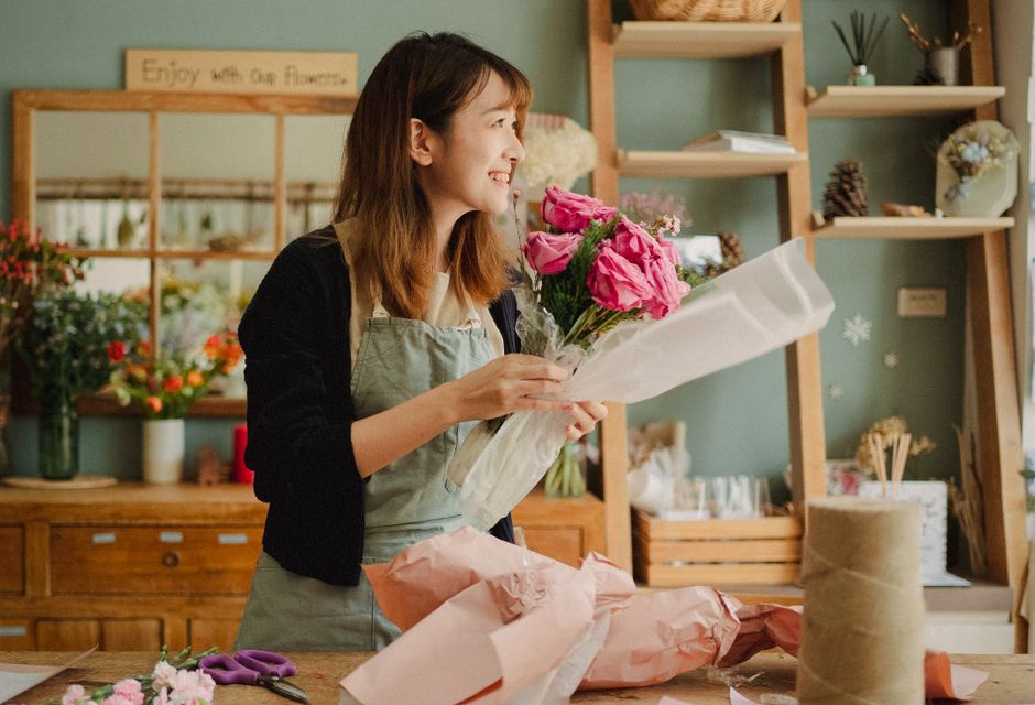 A smiling woman with shoulder-length brown hair, dressed in a black top and light-colored apron, holds a bouquet of vibrant pink roses and purple flowers wrapped in clear cellophane. She stands in a w