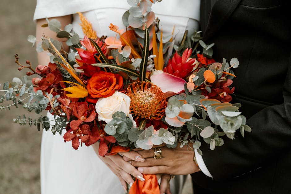 A close-up of a bridal bouquet featuring vibrant red, orange, and white flowers, including roses and protea, accented with silvery eucalyptus leaves. The bouquet is held by a bride and groom; the brid