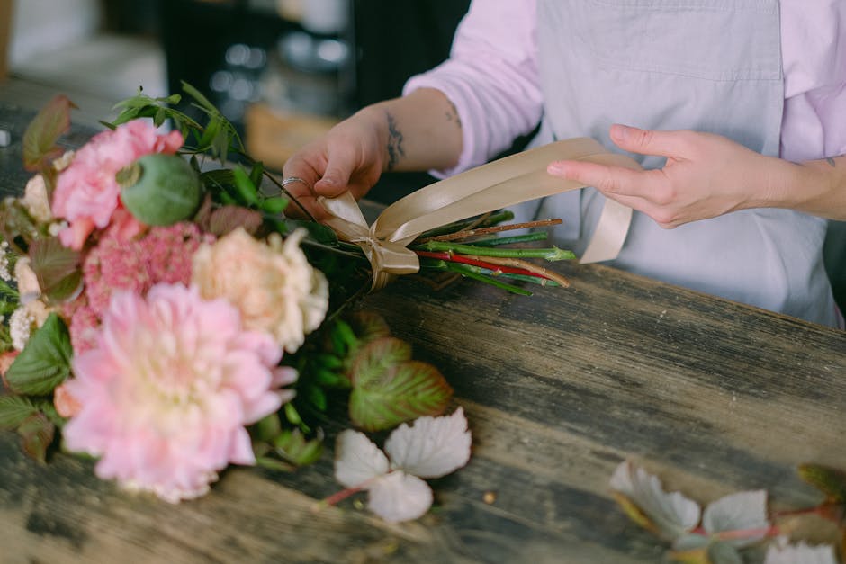 A florist dressed in a light pink shirt and white apron is carefully handling a bouquet of fresh flowers on a rustic wooden table. The bouquet features large, soft pink peonies, variegated leaves, and