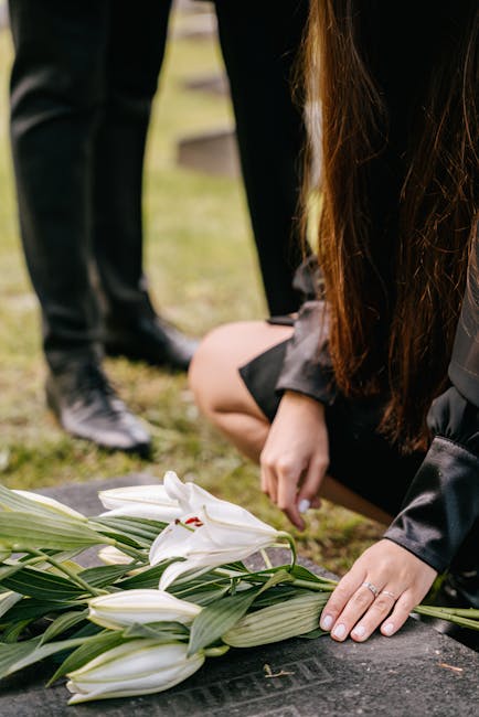 A close-up of a person kneeling at a grave, arranging a floral tribute with white lilies and greenery. The individual, dressed in dark clothing, gently places their hand on the flowers, which are wrap