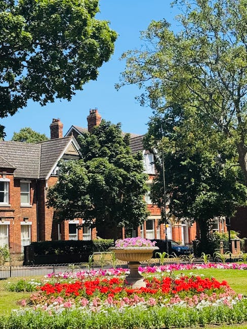 A vibrant urban park scene under a clear blue sky, featuring well-maintained flowerbeds with red, pink, and white blooms arranged in neat rows around a large stone urn filled with pink flowers. Tall,