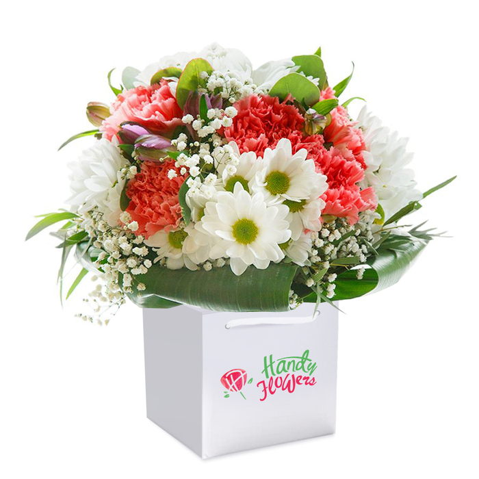 Presented in a spotless white gift bag against a seamless white background, this opulent Northolt bouquet feels like an embrace of spring's possibilities, artfully arranged with the skill of a seasoned neighbourhood florist. The eye is first drawn to plush carnations in an array of coral, peach, and red, their lavishly frilled petals cascading outward with tactile softness. Bright white daisy-like chrysanthemums punctuate the bouquet, each with a vibrant yellow-green center that brings cheer and harmony-much like a stroll through Northala Fields. A gentle network of white gypsophila nestles between the larger blooms, lending an airy, almost cloud-like texture. Sprigs of unopened buds, gently flushed with purple, are tucked thoughtfully on the left side, hinting at evolving beauty. Around the base, broad arcs of lush, glossy foliage cradle the blooms, mirroring the natural borders along Mandeville Road and unifying the arrangement's abundant shape. The gift bag's clean, modern lines are complemented by sleek white handles, designed for effortless gifting or local delivery in Northolt. A discreet logo signals local florist expertise. The bouquet radiates freshness, elegance, and ready-to-gift grace-suited to occasions from heartfelt birthdays to milestone anniversaries. This premium composition is a testament to the artistry found within Northolt's flourishing floral scene, inviting one to partake in the simple joy of giving and receiving flowers infused with local character.