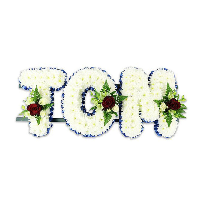 A bespoke memorial arrangement shapes the name TOM in large, three-dimensional letters, each letter densely enrobed in small, snow-white chrysanthemums that create a soft, tactile surface recalling a folded shroud of petals. The perimeter of every character is sharply outlined in royal blue ribbon-cool, vivid and precisely applied-so the letters read with clear definition against a stark white backdrop. Placed thoughtfully within each letter is a balanced floral spray: one rich, dark red rose serves as the visual and emotional centre, its velvety petals contrasted by smaller creamy-white spray roses and delicate green fern fronds that add air and texture. A discreet dark metal bar supports the lower section of the tribute, visible at the base and designed to hold the piece upright for viewing in a chapel, community hall, or outdoor service. Soft, diffused lighting highlights the chrysanthemum petals' layered structure and the subtle sheen of the ribbon, while a faint aroma of rose and green foliage lingers-an unobtrusive reminder of care and presence. This arrangement reads as both statement and solace: formal in composition, intimate in detail, and intended for moments of farewell. In the Northolt area-whether delivered to a service near Northala Fields or to a family home off Western Avenue-this floral name tribute expresses respect, steadiness, and a carefully measured tenderness from the hands of experienced arrangers.