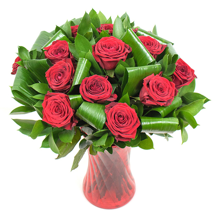 A carefully composed, high-impact bouquet featuring a dozen-plus deep crimson roses, each blossom fully open to reveal densely spiralled, velvety petals that suggest both freshness and refined luxury. The roses sit tightly clustered to form a plush, rounded mass, their rich red tones ranging from bright ruby highlights to darker, wine-tinged shadows where petals fold inward. Abundant dark green foliage fills the spaces between blooms: glossy, flat leaves-likely salal or ruscus-create a lush backdrop while larger, broad leaves styled like aspidistra have been rolled and curved into deliberate cylinders and loops, producing striking architectural accents that cradle the flowers and add sculptural depth. Those folded greens introduce a modern rhythm, their glossy textures contrasting with the soft matte of the rose petals. The assembly rests in a tall, elegant ruby-red glass vase, translucent with a subtle twisted or ribbed surface that throws back light in vertical flares and complements the floral tones. The vase narrows slightly at the neck then flares, lending balance to the dense crown of roses. Against a pure white background, the composition reads with dramatic clarity, every texture and contour made visible: the plush petal surfaces, the cool firmness of the leaves, and the warm, glassy glow of the vase. A seasoned florist in Northolt might present this as a romantic centrepiece ideal for anniversaries or Valentine moments, evoking the calm of an evening walk near Northolt High Street as much as the intimate warmth of a candlelit table.