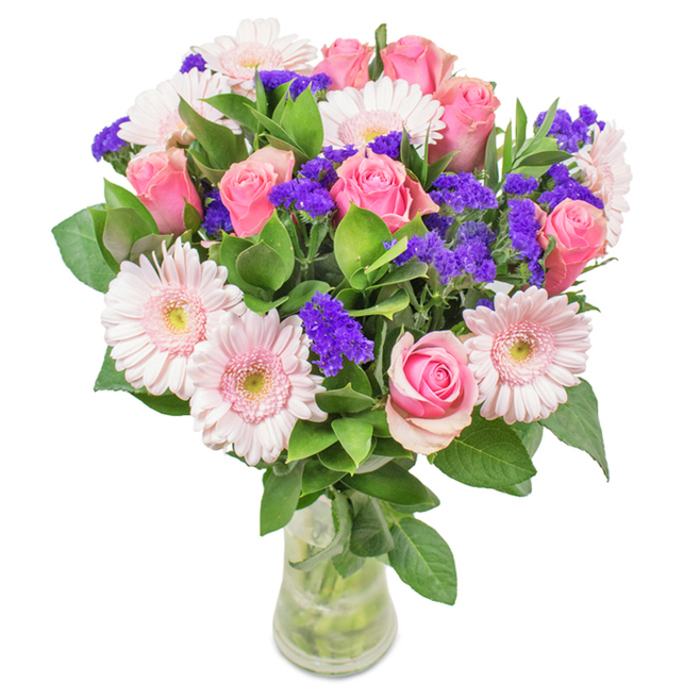 This enchanting floral arrangement, beautifully presented in a slender glass vase, captures the fresh, uplifting spirit of Northolt. At its heart, a medley of about nine elegant pink roses-some in tight buds, some just beginning to open-showcase timeless romance with their graceful spirals and layered, silky petals. They are surrounded by soft blush gerbera daisies, their generous, round heads and lighter centres lending a sense of gentle movement and airiness, reminiscent of blooms dancing in the spring breezes near Northala Fields. Sprigs of deep purple statice are woven throughout, providing striking contrast and a touch of wild texture. A rich layering of deep green foliage anchors the display, evoking both abundance and the enduring greenery of nearby parks. Subtle natural light reveals the intricate petal forms and the translucent stems within the clear water of the vase, reinforcing the notion of freshness and careful curation. This bouquet, crafted for joyous milestones or as a thoughtful gesture 'just because', effortlessly brings the refined artistry of a Northolt florist's touch into any home or workspace, making every recipient feel cherished and celebrated.