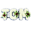 This image shows a classic sympathy flower tribute spelling out the name TOM in striking, three-dimensional capital letters, designed for a funeral or memorial service in Northolt. Each letter is completely covered with tightly packed white chrysanthemums, their petals forming a soft, plush surface that conveys purity and peace. A crisp royal blue satin ribbon is wrapped carefully around the edges of the T, O, and M, framing the white blooms with a formal, dignified border that stands out clearly against the plain white background. On the top left of the T, along the side curve of the O, and near the upper right of the M, elegant floral sprays break the uniformity of the chrysanthemums. In each spray, rich red roses open fully, their velvety petals creating a deep, emotional focal point. These are surrounded by smaller white or cream spray roses and slender white flowers reminiscent of freesia, which add lightness and a hint of movement. Lush green foliage-ferns and other leafy stems-fans out gently around the clusters, bringing natural contrast and depth to the design. The letters are arranged in a horizontal line on a narrow dark bar, providing a sturdy base so the tribute can be displayed along the side of a coffin, on a stand in a funeral home, or at a graveside in Northolt. The composition feels both traditional and heartfelt, a bespoke floral arrangement that allows family and friends to see Tom's name expressed in flowers as a final, loving message of remembrance and respect.