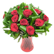 A lush, rounded bouquet of a dozen or more deep crimson roses fills the frame, each bloom in full, velvety perfection with petals that spiral tightly at the centre and unfurl into soft, satin edges. The red is almost wine-dark in places, intensified by even, soft light that reveals the delicate veining and the subtle, powdery sheen of fresh petals. Surrounding and cradling the roses is a generous mass of dark green foliage: flat, glossy leaves like salal or ruscus form a dense, verdant backdrop while broader, emerald-green leaves-reminiscent of aspidistra-are expertly folded, curled, and spiralled into architectural loops that peek between the blooms. Those sculpted leaf forms give the arrangement a modern, structural rhythm, their glossy surfaces catching small highlights against the petals' matte depth. The stems are gathered in a tall, slender ruby-red glass vase whose translucent body has a faint ribbed or twisted texture; vertical lines in the glass catch and reflect light, echoing the roses' upright stems and intensifying the composition's passionate reds. Set against a clean white background, the bouquet reads as a single statement of elegant romance and careful craftsmanship: tactile petals, cool polished leaves, and the glossy warmth of red glass. As a local florist might say when describing a gift for an anniversary or Valentine's Day in Northolt, this arrangement balances classic sentiment and contemporary design, a bouquet that feels as intimate as a handwritten note and as luxurious as an evening by Northala Fields at dusk.