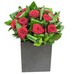A compact, modern arrangement of deep crimson roses gathered into a tight cluster at the heart of the design, their velvety petals unfurling in generous spirals that catch the light like polished silk. Around the roses, broad glossy green leaves have been carefully looped and curled into sculptural ribbons, creating a rhythmic pattern of movement and contrast against the blooms. The flowers sit low and confident in a matte black cube, the square silhouette lending a contemporary, urban edge that anchors the composition. The lighting is crisp and even, picking out subtle highlights on each petal and the sheen on the foliage so the crimson reads almost tactile, while the greens hold a cool, emerald clarity. Close to the arrangement you can imagine the clean scent of fresh leaves intertwining with the classic perfume of rose-bright, slightly green, and quietly uplifting. Texturally the petals feel silken and soft in contrast to the firmer, almost waxy surface of the leaves; visually the composition balances intensity and restraint. This bouquet reads as a deliberate statement of affection or admiration, suited to an anniversary, a thoughtful congratulations, or to lift an understated city space in and around Northolt village. The craftsmanship is visible in the folded foliage and the precise spacing of each bloom, a testament to an experienced hand arranging for simplicity and impact. Overall it is a study in refined romance and modern structure, a piece that feels as much like a small sculpture as it does like a gift, offering both visual drama and intimate warmth.